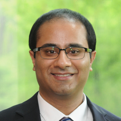A man with short black hair and glasses, wearing a dark suit, white shirt, and striped tie, smiles in front of a blurred green outdoor background.