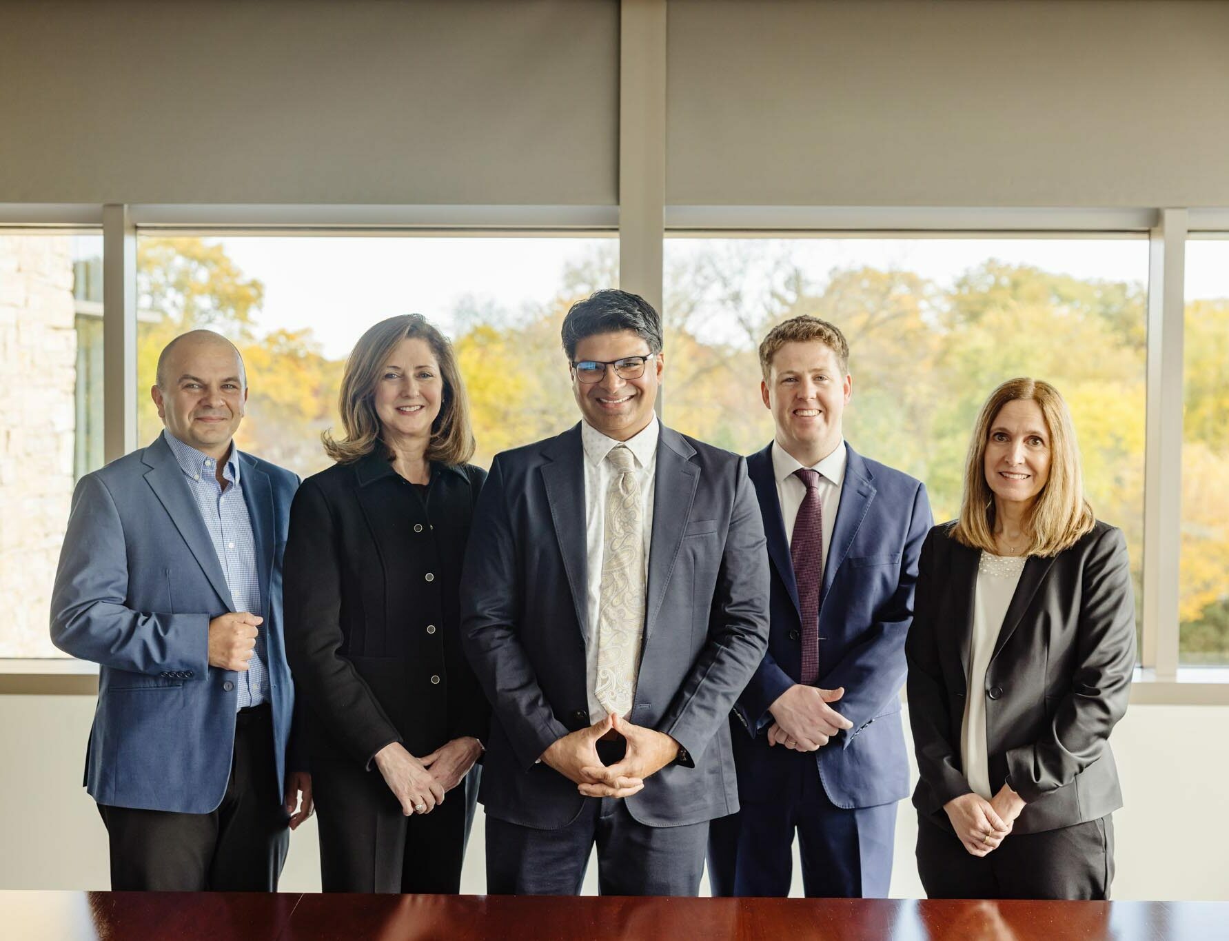 Five professionally dressed people stand side by side indoors in front of large windows with trees visible outside. They are smiling and facing the camera, suggesting a business or team setting.