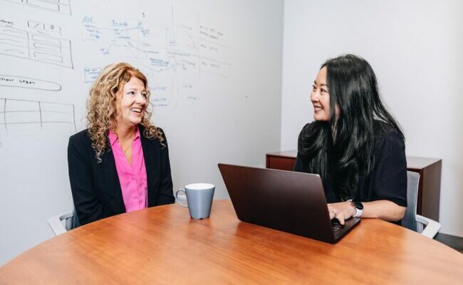 Two women sitting at desk talking with one on computer