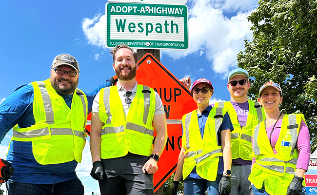 Five volunteers wearing neon yellow vests standing in front a green sign 