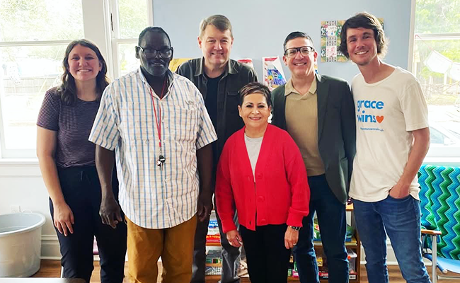 Six people are standing together in a group photo. There are two women and four men and they are all smiling.