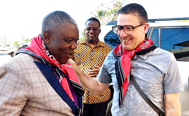 A man wearing a pinstripe tan blazer and a red bandanna is standing next to a man wearing eyeglasses, a red bandanna and gray t-shirt. Behind them is another man earing a button-up shirt.