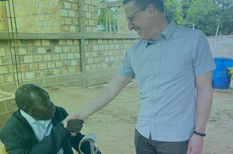 A man seated in a wheelchair is extending his hand and shaking the hand of a man who is standing across from him.