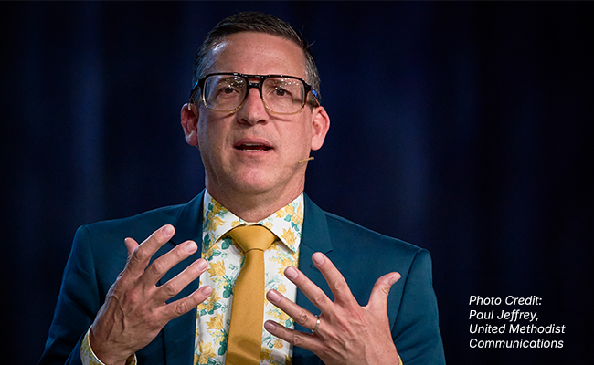 A young man wearing eyeglasses, a blue blazer, yellow tie and printed shirt is speaking and his hands are raised.