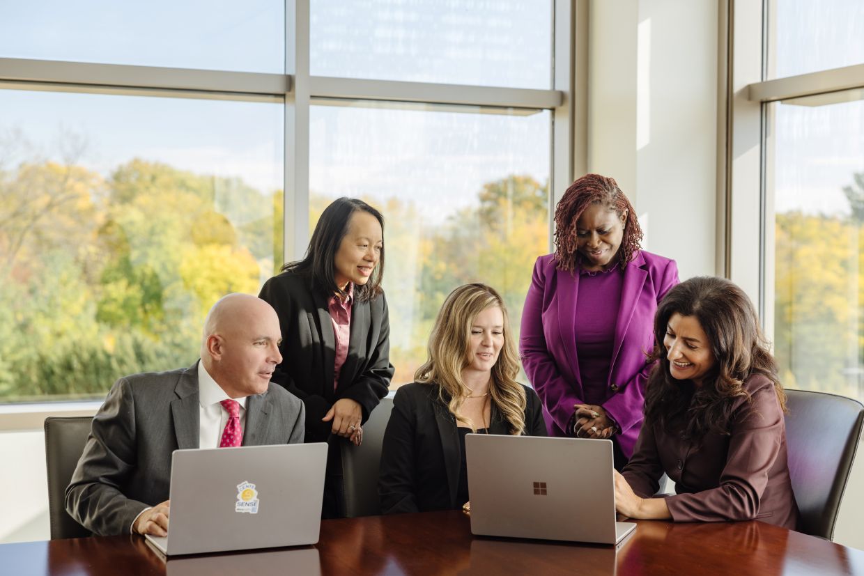 Five members of the investments team collaborating at a table with laptops. Three team members are seated and two are standing.
