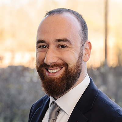 Portrait of Jon Morris smiling. He has a beard and is wearing a dark blazer, light collared shirt, and a dark tie.
