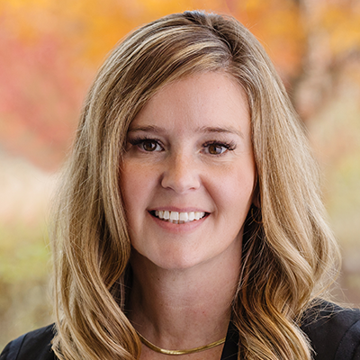 Portrait of Karen Manczk. She is smiling, has long blonde hair, wearing a gold necklace and black jacket.