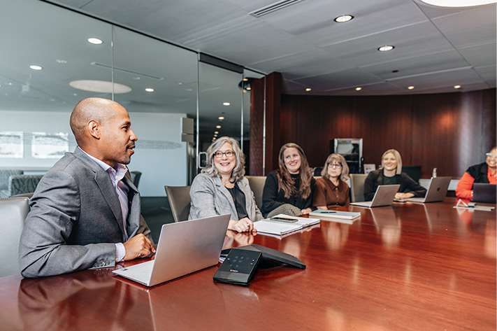 A man in a gray suit is sitting at a table with a laptop. She is facing five women. The women are seated at the table and smiling.