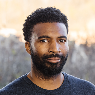 Portrait of Micah Burkley smiling. He has a beard, dark short hair and is wearing a black shirt.