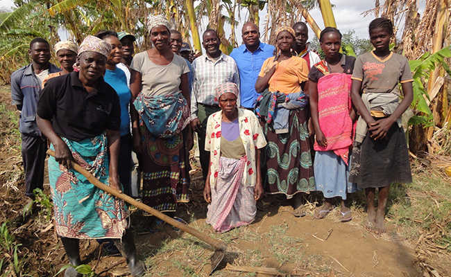 A group of fourteen people are standing together. One woman in the front is holding a garden hoe.