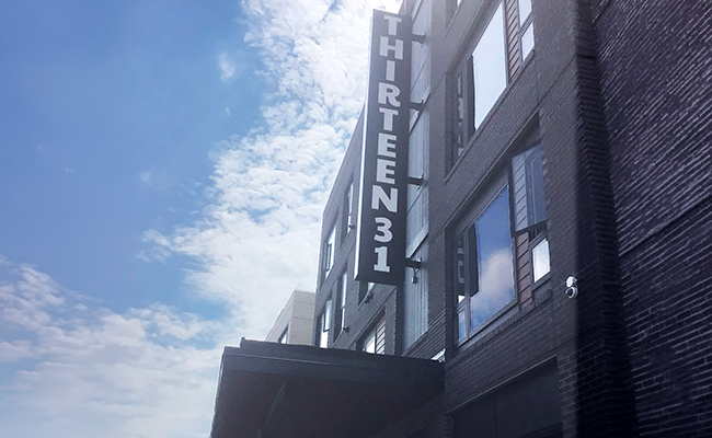 The outside of a dark brick building with a vertical black sign with white lettering 