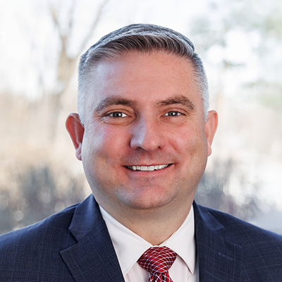 Portrait of Trent Sparrow smiling. He's wearing a black blazer, white collared shirt and a red tie.