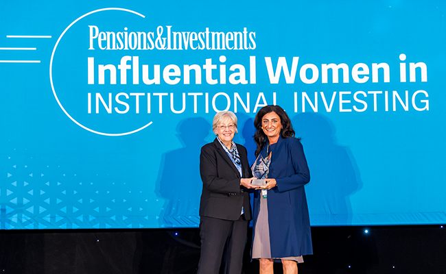Two woman standing next to each other and holding an award. They are standing in front a a blue sign with white lettering reading Pensions & Investments Influential Women in Institutional Investing.