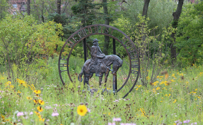 A metal statue of John Wesley on a horse outdoors surrounded by trees, flowers and grass.