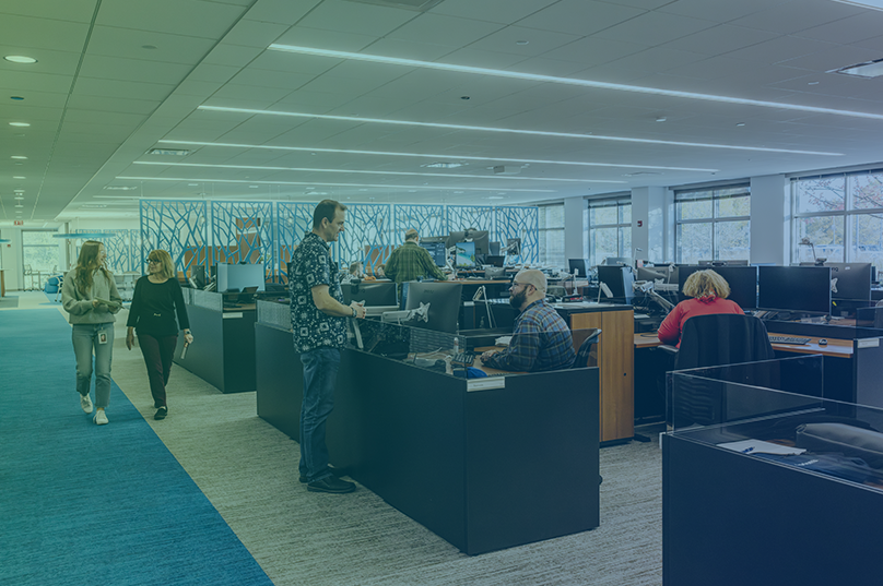 Open office floor plan with cubicles. Some employees are standing and some employees are seated.